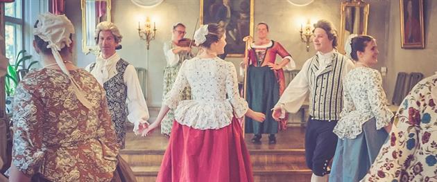 Personer som dansar 1700-tals dans i tidstypiska kläder i vacker miljö.