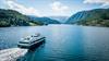 Fjordcruisebåten Fjorddrott seiler innover Hardangerfjorden med snødekte fjell i bakgrunnen, sett frå drone.
