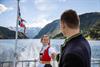 Par i bunad held kvarandre i handa på akterdekket under fjordcruise på Hardangerfjorden, norsk flagg i bakgrunnen.