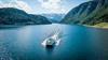 Fjordcruisebåten på Hardangerfjorden sett frå drone forfra, med blått vatn og snødekte fjell på begge sider.