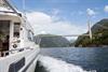 Hardanger Fjordcruise under an impressive bridge in the Hardangerfjord, surrounded by mountains and a blue sky.
