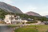 A large wooden white guesthouse, surrounded by high mountains and greenery, during sunset.