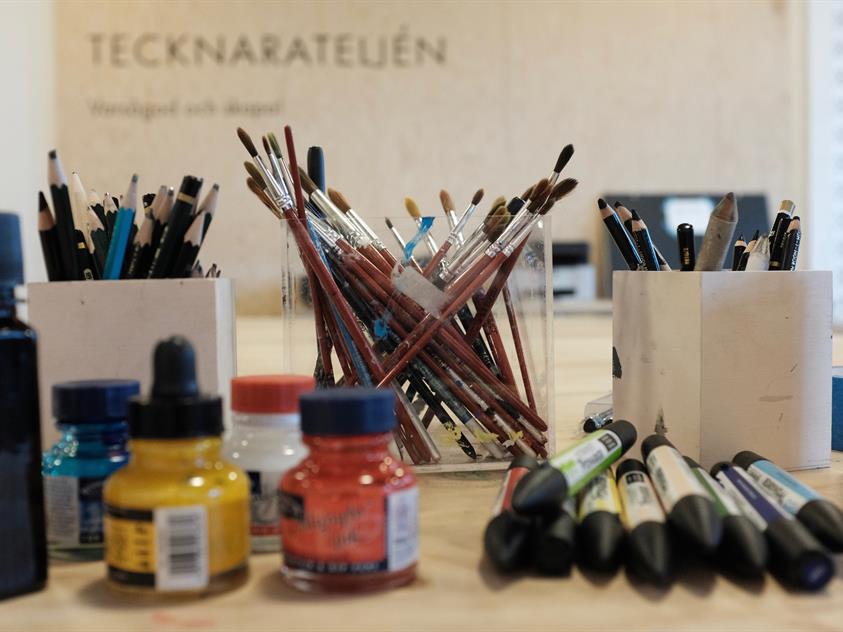 Bilden visar olika konstnärsmaterial såsom penslar, kolpennor och burkar med flytande tusch.